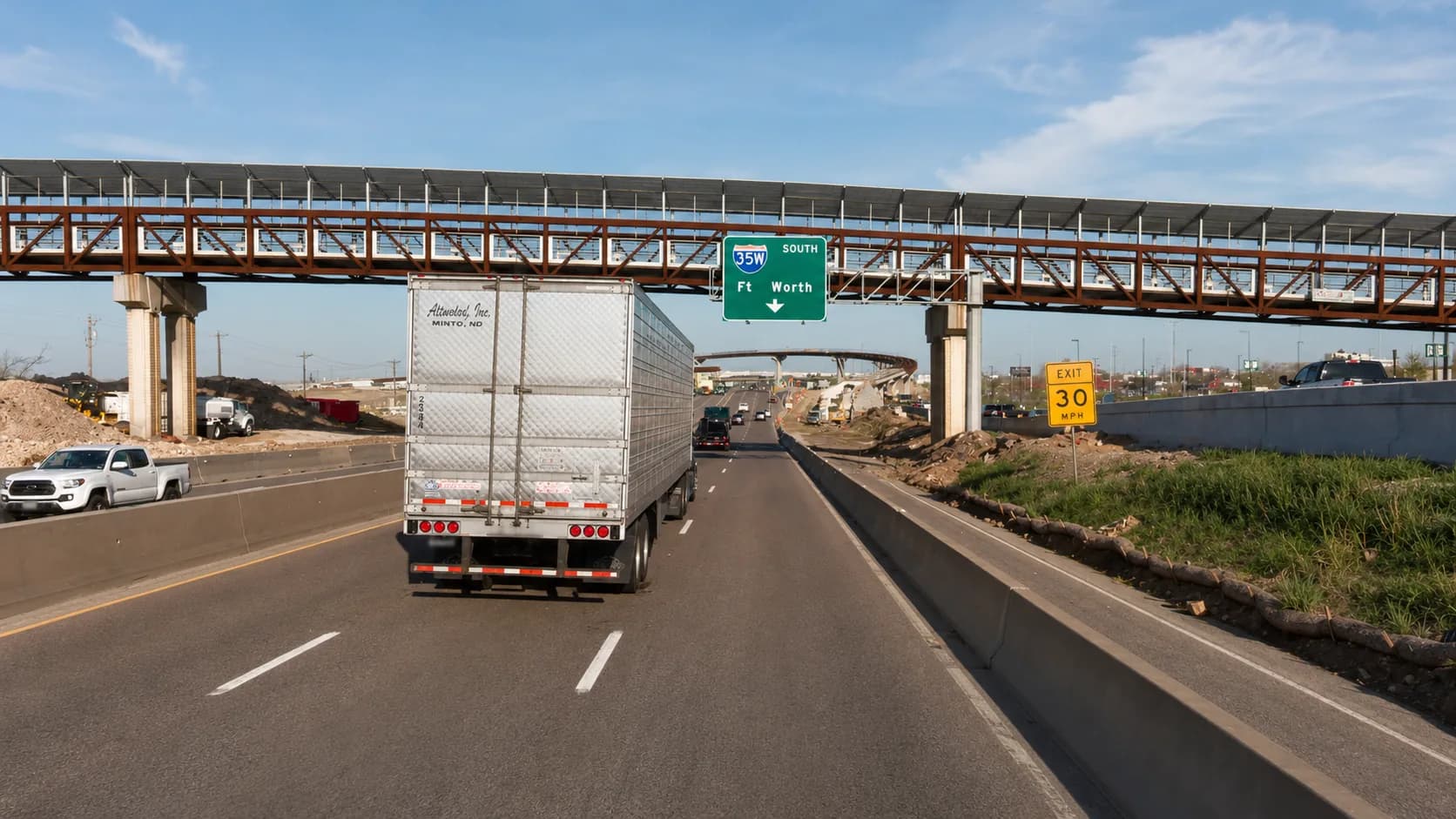 Interstate 35E through Carrollton, Texas, with an 18-wheeler hauling a refrigerated trailer approaching a highway overpass and overhead signage in a construction zone under a clear Texas sky