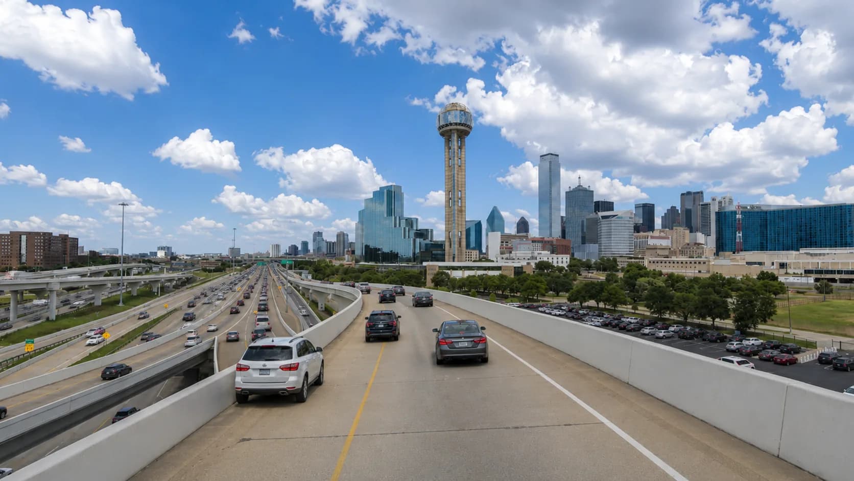 Passenger vehicles backed up on a Dallas freeway near the downtown Dallas skyline and the Interstate 35E and Interstate 30 interchange under a bright Texas sky