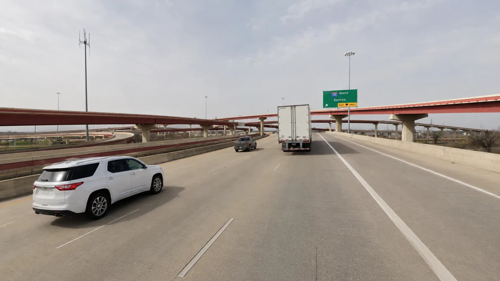 Interstate 35 through Denton, Texas at the Interstate 35E and Interstate 35W split, with a box truck and passenger vehicles beneath the rebuilt red flyover ramps under a hazy Texas sky