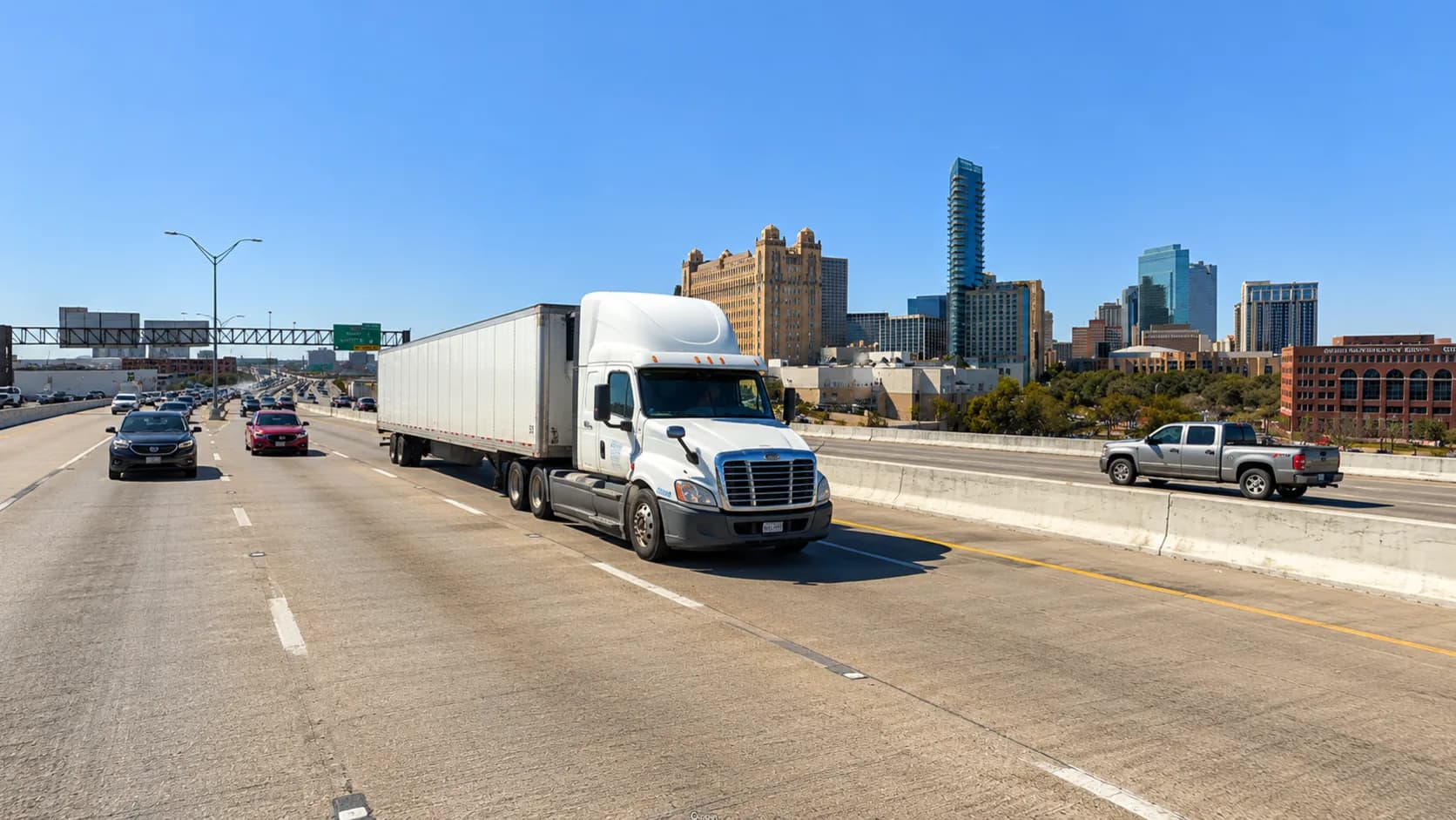 Interstate 35W approaching the downtown Fort Worth, Texas skyline, with a white 18-wheeler and passenger vehicles on the multilane freeway under a bright Texas sky