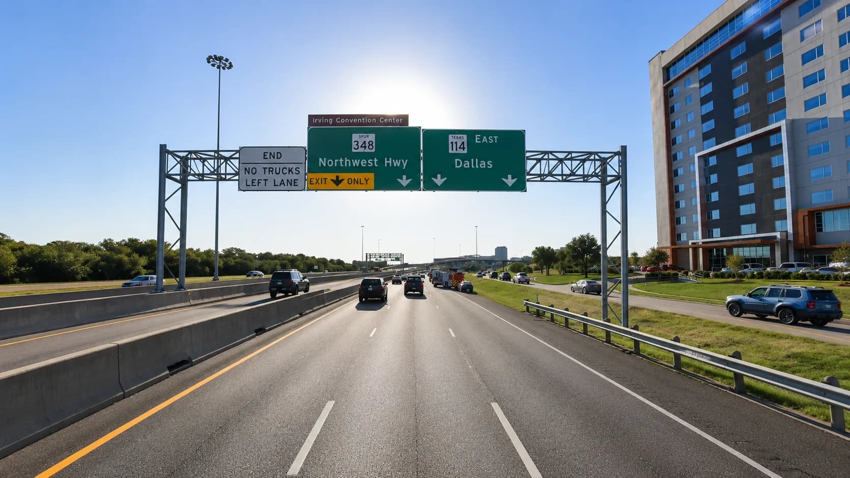 State Highway 114 east toward Dallas through Irving, Texas, with an 18-wheeler and passenger vehicles approaching the Spur 348 / Northwest Highway interchange near Las Colinas and the Irving Convention Center under a bright Texas sky