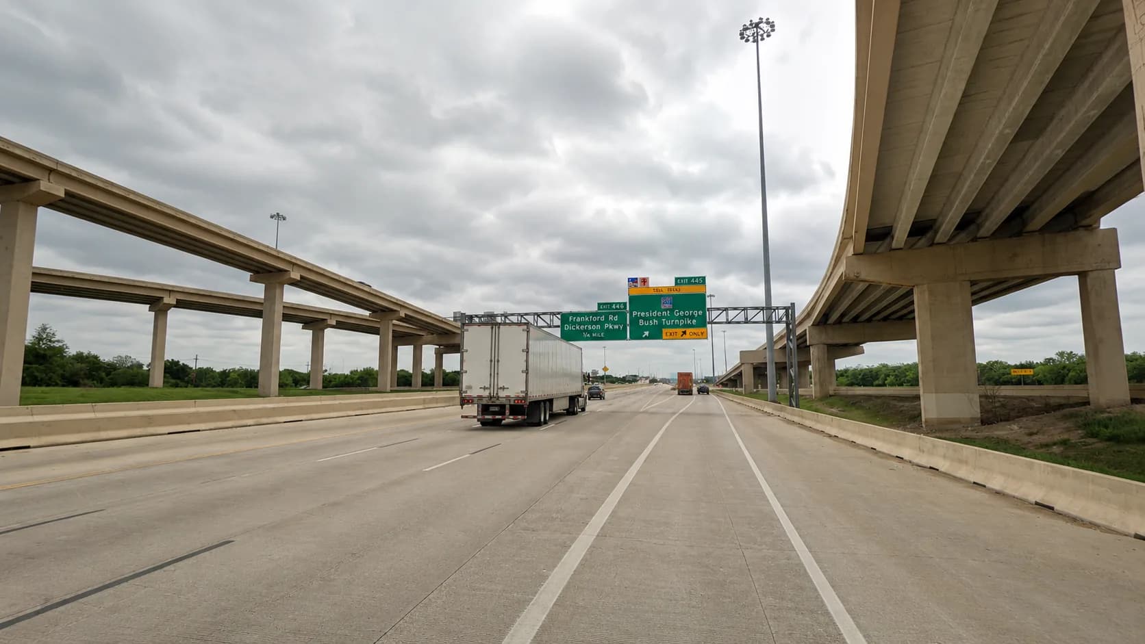 Interstate 35E through Lewisville, Texas, near the Sam Rayburn Tollway interchange, with an 18-wheeler and passenger vehicles beneath the concrete flyover ramps under an overcast Texas sky