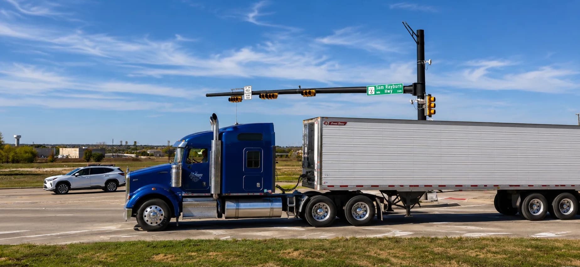 A blue 18-wheeler hauling a white trailer through a major intersection on the Sam Rayburn Highway / State Highway 121 corridor in Melissa, Texas, alongside a passenger vehicle under a bright Texas sky