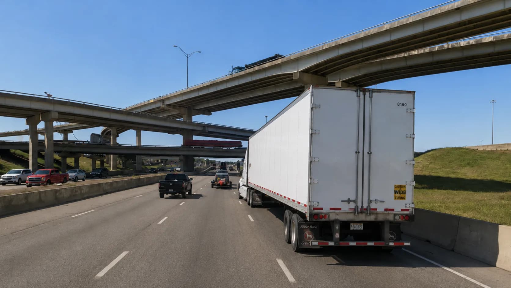 Interstate 635, the LBJ Freeway, through Mesquite, Texas, with a white 18-wheeler and passenger vehicles approaching the stack interchange flyovers under a bright Texas sky